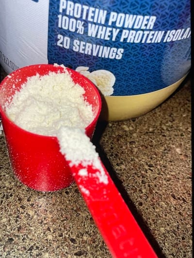 A close-up shot shows a red scoop filled with white protein powder, resting on a speckled countertop. Behind the scoop, a portion of a large white and blue container of protein powder is visible, with text indicating "PROTEIN POWDER 100% WHEY PROTEIN ISOLATE 20 SERVINGS." The background is a speckled brown countertop. The image does not contain any information about the city of Neubrandenburg, Germany.
