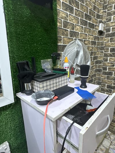 A barber's workstation with an open drawer revealing hair clippers and a power adapter inside. On top of the drawer are a black-handled brush, a blue comb, various bottles, and a tissue box. A gray cape is draped beside these items. The workspace is set against a brick-patterned wall and artificial grass. A white-framed mirror is visible at the bottom of the frame.
