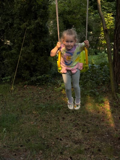 A young child is swinging on a yellow swing set in a backyard. The child has blonde hair and is wearing a tie-dye t-shirt, grey leggings, and white Crocs with various charms. The child's arms are wrapped around the ropes of the swing, and they are looking towards the camera with a slight smile. The swing set is positioned in front of a backdrop of lush green trees and foliage, with a hint of a wooden fence visible in the background. The ground is covered in grass and scattered pinecones, with patches of sunlight breaking through the trees, suggesting it is a sunny day. The overall scene evokes a sense of childhood joy and outdoor play. The image appears to have been taken in the late afternoon, given the warm lighting and shadows.