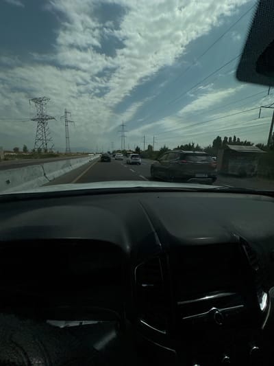 The image shows a view from inside a car driving on a highway. The highway has multiple lanes and is divided by a concrete barrier. Several cars are visible on the road, including a dark SUV in the right lane with a license plate that appears to read "UJ045JB". There are large power line towers on the left side of the highway. The sky is cloudy with patches of blue. Trees and some low buildings are visible on the right side of the road. The dashboard of the car is visible in the foreground, partially obscuring the view. The image appears to have been taken in the city of Istiqlol, Uzbekistan, based on the presence of a license plate with Cyrillic characters, commonly used in Uzbekistan.