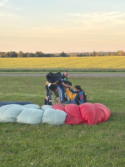 In the city of Limbaži, Latvia, at what appears to be a skydiving drop zone during sunset, three individuals in skydiving gear are interacting on a grassy field. In the foreground, three large, deflated parachutes are laid out on the grass. Two are light blue and white, and one is bright red. In the middle ground, two individuals are bending down, seemingly assisting a third individual who is sitting on the ground. The person on the ground is wearing a blue jumpsuit and a helmet. Another person, also in a blue jumpsuit, is standing and reaching towards the seated individual. A third person, wearing a black backpack and a black and orange outfit, is standing and appears to be supervising or assisting. The background features a flat, grassy field, a line of trees, and a large, yellow, cultivated field, all under a clear sky with soft, warm sunset lighting.