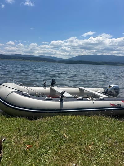 This is a photograph taken on a sunny day in Tsigov Chark, Bulgaria. The scene features an inflatable boat with a Honda outboard motor docked on the grassy shore of a lake. The boat is white with blue and red accents, and it has fishing rod holders and a depth finder mounted on it. The lake's water is rippling, and in the background, rolling hills covered with trees and vegetation stretch towards the horizon under a bright blue sky with scattered white clouds. The foreground consists of green grass with scattered wildflowers.