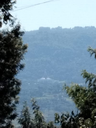 The image displays a panoramic landscape captured from an elevated viewpoint in Subukia ward, Kenya, showing the National Shrine of Mary Help of Christians situated within a valley. The foreground features dark, dense tree foliage partially framing the scene from the left and right sides. The midground reveals a lush, green valley containing scattered vegetation and a prominent white building characterized by two distinct, light-colored conical or spired roofs. This structure, identifiable as the Subukia Shrine, is surrounded by green terrain. The background consists of hazy, densely forested hills or mountains under a clear, light blue sky. A thin, dark linear object, possibly a utility wire, traverses the upper portion of the sky. No individuals or specific actions are discernible.