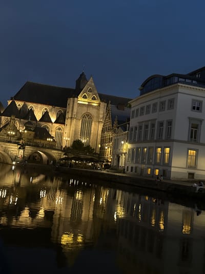 An evening shot shows a canal with reflections of illuminated buildings on its surface. On the left, a large Gothic cathedral, likely St. Bavo's Cathedral in Ghent, Belgium, is visible with its arched windows and pointed spires. Adjacent to it, on the right, is a more modern, white building with multiple rectangular windows. The sky is a deep blue, indicating it is twilight or nighttime. Several people are seated on the canal's edge, and others are dining at outdoor tables, suggesting a public space or promenade. The overall scene depicts a European city at night, characterized by historic architecture and a calm waterway.