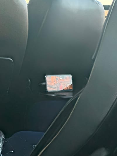 An eye-level shot reveals the interior of a vehicle, likely a car. A dark, padded headrest occupies the left side of the frame, while a portion of a dark dashboard or console stretches across the top. Adhered to the back of the headrest is a smartphone in a clear case, appearing to be charging via a cable. The phone screen displays a blurry image, possibly a video or live feed. A navy blue portion of a seat is visible in the bottom right.
