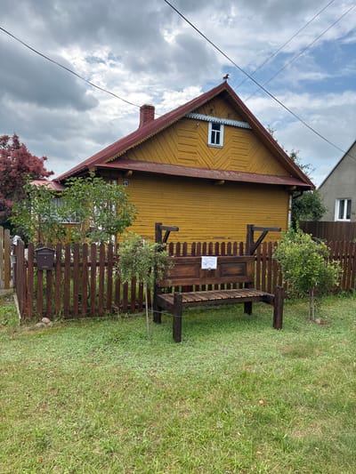 The image captures a scene in the village of Trześcianka, Poland. A weathered, mustard-yellow wooden house with a red roof and decorative eaves is the prominent structure. A red-brick chimney rises from the left side of the roof, and a small, white-paned window is visible on the gable end, topped by a decorative weather vane shaped like a horse. A dark brown wooden fence surrounds a grassy yard in front of the house. In the yard, a rustic dark brown wooden bench sits on the grass. A mailbox is attached to the fence near a leafy green tree. The sky is overcast with dramatic clouds.