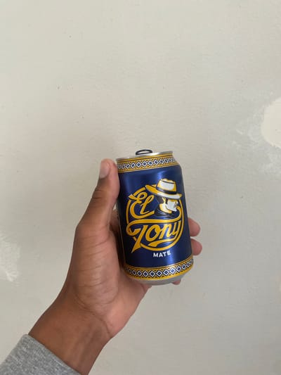 A human hand, with a dark skin tone, is holding a beverage can against a plain, light-colored wall. The hand is positioned towards the right side of the frame, holding the can horizontally. The can is dark blue with prominent yellow branding.

The main text visible on the can is "EL TONY," written in a stylized yellow script. Above this text is a logo featuring a character wearing a hat, also rendered in yellow and white tones. Below the main text, oriented vertically, is the word "MATE" in smaller yellow letters. The top and bottom edges of the can's blue label are adorned with a repeating geometric diamond pattern in black and white or silver. The can's opening tab is lifted, indicating it has been opened.

The background is an off-white or light gray wall with a slightly textured surface. There are some faint, lighter patches and subtle marks visible on the wall, particularly in the upper right corner. Part of a gray sleeve is visible on the person's wrist. The setting is simple and focuses on the interaction with the beverage. The context of the image is Villeurbanne, France.