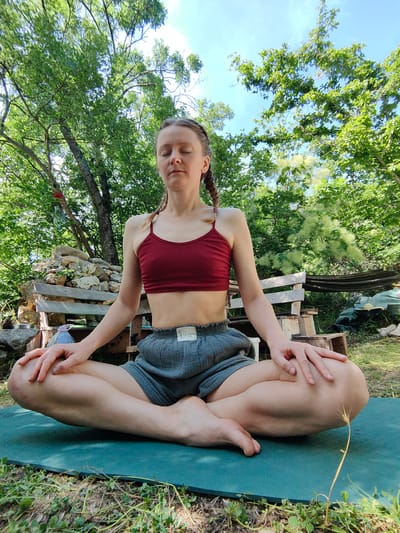 A woman is meditating outdoors on a yoga mat. She is sitting in a cross-legged position, with her eyes closed and her hands resting on her knees. She is wearing a dark red sports bra and grey shorts. The background shows a lush green environment with trees and a blue sky. There is a rustic wooden bench and a pile of rocks behind her. The scene appears to be set in a natural, serene location, possibly a garden or a park. The time of day seems to be daytime, likely afternoon, judging by the sunlight and shadows. The overall mood is peaceful and contemplative. Visible text on her shorts reads "La Luna".