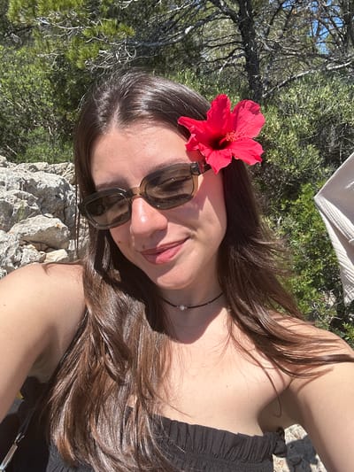 A young woman lies on a light-colored rock formation, likely limestone, surrounded by green shrubs and trees, possibly in the municipality of Calvià, Spain. She wears dark sunglasses and a red hibiscus flower is pinned behind her glasses. Her hair is long and brown. She is wearing a black top. A small pearl necklace can be seen on her neck. The photo is taken in bright daylight.