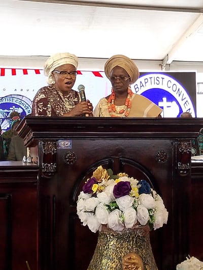 This photograph captures a formal event, likely a Baptist Convention in Abuja, Nigeria, as evidenced by the partial text "BAPTIST CONVEN" and "ABUJA" displayed on a large screen in the background. Two women, dressed in traditional Nigerian attire including elaborate headwraps (geles), stand behind a richly carved, dark wooden podium or pulpit.
The woman on the left is actively speaking into a microphone, her mouth open as if mid-sentence. She wears glasses and a patterned brown and cream outfit with a cream-colored headwrap. Beside her, the woman on the right stands attentively. She also wears glasses, a light gold-colored outfit with a matching headwrap, and a distinctive coral bead necklace with large, long coral earrings.
In front of the podium, a decorative floral arrangement in a textured gold vase adds a touch of elegance, featuring white, purple, yellow, and blue artificial flowers. The setting appears to be an indoor or covered outdoor venue, possibly under a large tent, indicated by the white ceiling structure visible at the top of the frame. The event conveys a sense of public address or religious gathering in Dushepe, Nigeria.