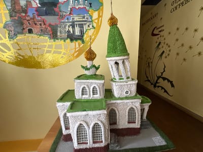 A miniature model of an Orthodox church with green roofs and white walls is displayed on a wooden surface in front of a yellow wall. The model has multiple levels and domes, with arched windows and intricate details. Behind the model, a large, colorful circular map-like image is mounted on the wall, featuring various buildings and a statue. The wall also has decorative elements, including dandelion motifs and text in what appears to be Cyrillic script. The overall setting suggests an indoor display, possibly in a museum or cultural center. The lighting is even and appears to be artificial, indicating it's not a specific time of day. There is no apparent activity or event taking place, as the scene focuses on the static display.
Visible text includes "МАСТЕРСКАЯ" (Workshop) and "ОТДЫХА СОГРЕВА" (Rest Warming) on the wall, along with smaller phrases and what looks like a QR code on the base of the model. The reference to "Ветрынскі сельскі Савет, Belarus" provides a geographical context for the display.