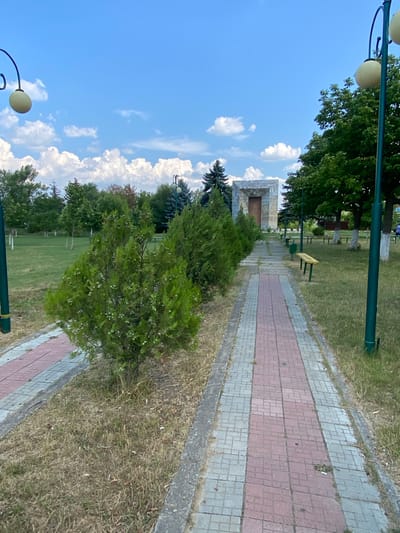 The image depicts a paved walkway in a park-like setting. The walkway is composed of red and grey square tiles, bordered by concrete. On the left side of the walkway, there are trimmed green bushes, and further back, a grassy area with scattered trees. On the right side of the walkway, there is also a grassy area with trees, and several park benches are visible. In the background, a large, stone monument or structure with a wooden door is situated. The sky is blue with scattered white clouds. Two dark green lampposts with spherical light fixtures are present in the foreground and mid-ground. The overall impression is of a public outdoor space, potentially a memorial or a section of a larger park. The context provided specifies the location as Ruseștii Noi, Moldova, suggesting this is a scene from that area. No people are clearly identifiable as the main subjects or actively engaged in interactions. The environment appears well-maintained but with some signs of natural wear, such as dry grass.