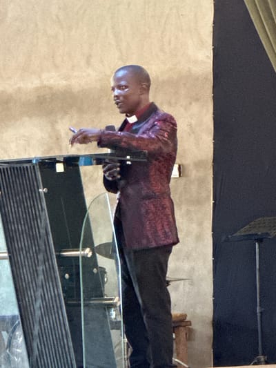 A dark-skinned man wearing a white clerical collar, a maroon sequined jacket, and dark pants stands behind a glass lectern. He is holding a pen in his right hand and appears to be speaking. The background consists of a textured, light-colored wall on the left and a dark curtain on the right. A music stand is visible in the background. The scene suggests a religious or public speaking event. The location is identified as Kufana, Nigeria.