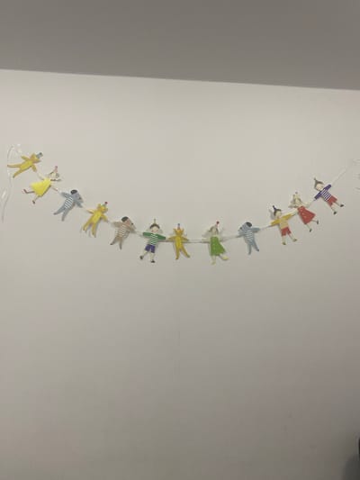 This media file shows a garland of cartoon figures, resembling children and animals, hanging from a white wall. The figures are depicted in various poses, each attached to the next by a thin string. They are colorful and appear to be made of paper or a similar lightweight material.
The figures include what seem to be people dressed in different colored outfits, such as a person in a purple and white striped shirt and red pants, another in a red dress, and one in a yellow shirt and red pants. There are also animal figures, some appearing to be bears or dogs, in yellow, brown, and blue. Several figures are wearing party hats, suggesting a celebratory or festive context.
The garland is suspended vertically, extending downwards from the top of the frame. The background is a plain white wall, with a slight textural detail visible. To the left of the garland, there is a distinct vertical line separating the white wall from a darker, grayish area, suggesting a corner or a transition between two surfaces or rooms.
No specific time of day or weather conditions can be inferred from the image. The overall impression is one of a playful and cheerful decoration. There is no visible text in the media file. The location context of Мирный, Russia, provides a geographical setting for this image.