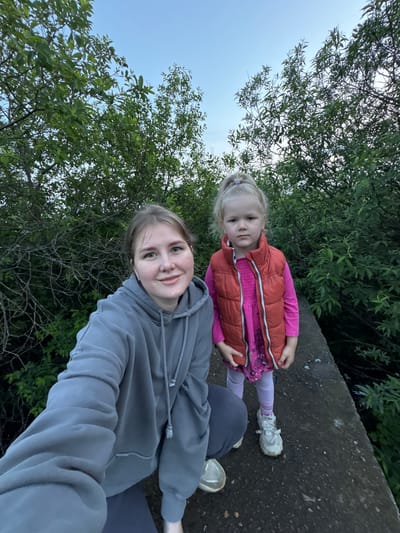 An outdoor, eye-level, full-shot selfie features two females in a wooded area. The woman on the left, in the foreground, is wearing a grey hooded sweatshirt and has her arm extended to take the photo. She has light skin, blue eyes, and light brown hair pulled back. She is smiling and looking at the camera. Next to her, to the right and slightly behind, is a young girl with blonde hair pulled into a high ponytail, wearing a pink long-sleeved shirt, a bright orange vest, light pink leggings, and white athletic shoes. She is looking directly at the camera with a neutral expression. They are standing on a concrete structure that appears to be a path or a bridge. The background is filled with dense green foliage and trees. The sky above is a pale blue, indicating either early morning or late evening light. The overall scene suggests a casual outdoor outing in a natural setting, likely near Горный, Russia, given the contextual information.