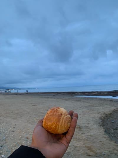 A hand holds a golden-brown pastry against a backdrop of a sandy beach and overcast sky. The sea is visible in the distance, with faint lights suggesting a town or city along the shoreline. The pastry features a swirling pattern, indicating layers within. The overall scene has a calm and somewhat muted color palette.