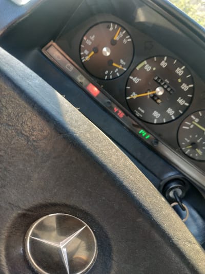 A close-up, angled shot shows the interior of a vehicle, focusing on the dashboard and steering wheel. The dashboard features several gauges including a speedometer displaying 31708 km, a tachometer, and a fuel gauge. Indicator lights are visible, with a red light illuminated and a green light displaying "PL1". The steering wheel, which has a silver Mercedes-Benz emblem, is partially visible in the foreground. The environment appears to be outdoors, with sunlight casting shadows. The scene is consistent with a vehicle parked on a street in Cherepovets, Russia, given the context provided.