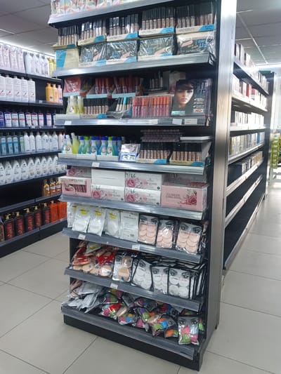 This image depicts an interior view of a retail establishment located in Kawaji, Nigeria, specifically an aisle stocked with personal care, beauty, and cosmetic products. The scene is dominated by multiple black metal shelving units.
On the left-hand side, a tall shelving unit displays an array of personal care items. The upper shelves feature white and colored bottles, including "Dr. Teal's" branded products, followed by numerous white and cream-colored bottles primarily from the "Simple" and "Dove" brands, likely containing shampoos, conditioners, or lotions. The lowest visible shelf on this unit holds darker, ornate bottles, possibly bath oils or specialty shower gels.
The central shelving unit, positioned prominently, focuses on beauty and cosmetic products. The top two shelves present multiple boxes of "WARDABEAUTY" concealers or foundations, labeled as "6 Colors, 48 Pcs." Below these are packages of "FIT ME Perfect Concealer" featuring a woman's image, alongside more "WARDABEAUTY" items. The mid-level shelves display various smaller cosmetic items, including liquid lip products, and bottles containing brightly colored liquids such as yellow, green, and blue, possibly cleaning solutions or liquid soaps. Further down, boxes of "LIP GLOSS" and "ULTIMATE LONG STAY" from the "Daisy Rose" brand, characterized by floral designs, are neatly arranged. The lowest shelves of this unit are filled with packaged "LEMON BEAUTY" brand makeup sponges and blenders in various colors and shapes, with visible price tags indicating "1,000" and "7,300."
To the right, another shelving unit extends down the aisle, presenting a similar inventory of products, though some of its shelves are sparsely stocked or empty towards the background. The floor is light-colored tile, and the ceiling features grid panels with integrated fluorescent lighting. No individuals are visible within the frame.