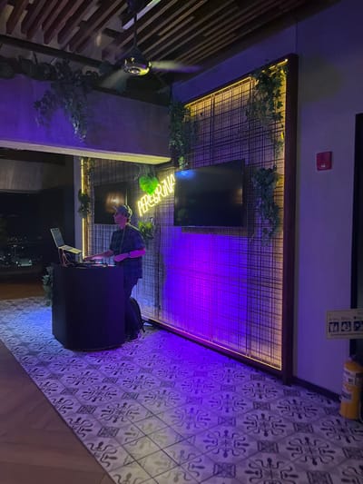The image depicts an interior scene of an entertainment venue named "Peregrina" in Floridablanca, Colombia, during nighttime. A male individual, wearing a plaid shirt, dark pants, and a baseball cap, stands behind a dark, hexagonal DJ booth. He is actively operating a laptop and a DJ mixer on the booth's surface. The floor beneath is tiled with a repeating geometric pattern in shades of white and gray.
The background features a prominent wall section composed of dark metal grid panels, framed by vertical and horizontal yellow LED strip lighting. Artificial ivy is draped over the grid and along the top edge. A green neon sign spelling "PEREGRINA" is mounted centrally on this grid wall. Two black, rectangular televisions are also wall-mounted, one partially obscured by the DJ, and the other fully visible. The area is illuminated by pervasive purple ambient light emanating from sources beyond the frame and reflecting strongly on the grid wall and floor.
Above, a dark metallic ceiling fan with four blades rotates, visibly emitting a fine mist. The ceiling itself features exposed wooden beams. To the right, a plain white wall section includes a red wall-mounted fire alarm and a yellow rectangular informational sign. A yellow cylindrical fire extinguisher rests on the floor at the base of this wall. To the left, beyond the DJ setup, a dark exterior is visible, punctuated by faint, distant city lights, indicating an elevated or panoramic location.