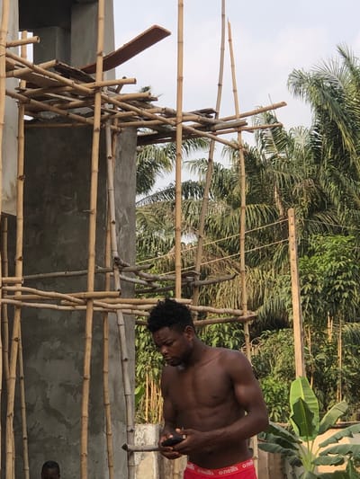 An adult male stands shirtless, facing forward and looking down at a black smartphone held in both hands. He has a dark complexion, a muscular build, and short dark curly hair. He wears red boxer briefs with "O'NEILLS" branding. To his left, a concrete building structure is under construction, surrounded by extensive bamboo scaffolding secured with lashings; wooden planks are positioned across parts of the scaffolding. A small child's head is partially visible in the lower-left corner, obscured by the concrete. In the background, dense tropical vegetation, including numerous palm trees and a banana plant, is visible under a bright sky. A wooden utility pole with visible wires stands on the right. The scene depicts construction activity within a vegetated area in Enugwu-Ukwu, Nigeria.