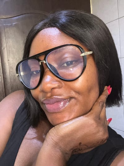 A close-up image depicts a single adult female of dark complexion, positioned in an indoor setting within the city of Dzhidu, Nigeria. The exact location within Dzhidu is not discernible from the image. The subject has short, dark hair styled in a bob and wears large, black-framed aviator-style glasses with clear lenses and gold-toned accents. Her lips are glossy and slightly parted in a faint smile. She is wearing a black sleeveless garment. Her right hand is raised, with the palm facing inwards, resting beneath her chin and jawline. The fingernails on her right hand are painted with red polish. A faint dark, intricate marking or design is visible on the dorsal surface of her right hand and wrist. The background on the left features a dark, possibly wooden, vertical surface, while the right background shows light-colored square tiles. Lighting appears to be even, illuminating the subject's face directly.