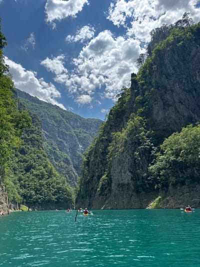 This is a vertical, slightly tilted, wide-angle shot taken in Bezuje, Montenegro. The scene depicts a serene mountain lake with a sheer, tree-lined cliff face on the left and a vast expanse of turquoise water on the right. A clear blue sky with fluffy white clouds is visible in the upper left portion of the frame.

Several people are engaged in kayaking on the water. In the foreground, a kayak with a person in a red and yellow life vest is clearly visible. Further in the distance, two more kayaks can be seen, suggesting a small group activity. The sun is shining, casting bright highlights on the water and illuminating the lush green trees clinging to the mountainside. The overall mood of the image is peaceful and adventurous, capturing the beauty of the natural landscape and the enjoyment of outdoor recreation. There is no visible text in the image.