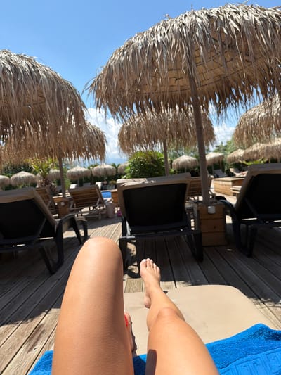 A person's legs are positioned on a lounge chair by a swimming pool, with a blue towel underneath. The foreground shows the person's tanned legs, with one foot visible at the top right. In the background, there are several thatched umbrellas providing shade over lounge chairs. The setting appears to be a resort or a private outdoor area, with wooden decking and lush greenery. The sky is clear blue with a few white clouds. The location is not explicitly identifiable as being in Plovdiv, Bulgaria.