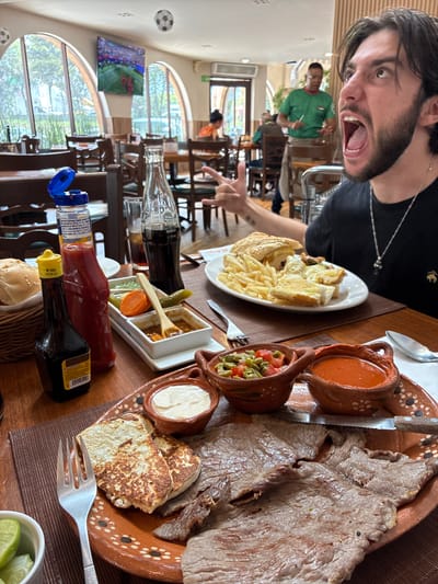 A young adult male with dark hair and a beard is captured in a close-up shot, his mouth wide open in a shout or exclamation. He is wearing a black t-shirt and a silver necklace. In the background, a restaurant setting is visible, with wooden tables and chairs, arched windows, and a television displaying a soccer match. A waiter in a green shirt stands behind the male. The foreground features a meal on a table, including grilled meat, cheese, sauces, and a side of fries. The scene appears to be in Cuautitlán Izcalli, Mexico.