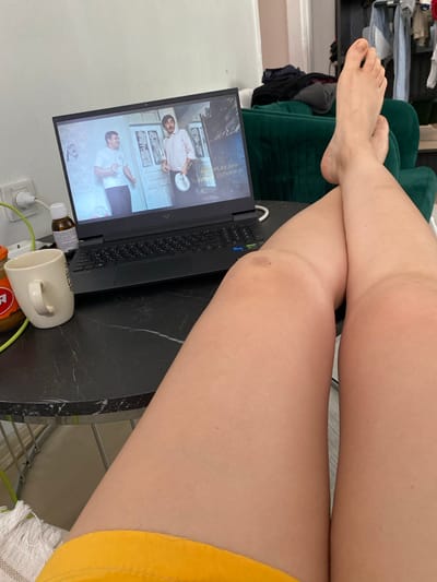 A person's legs are positioned in the foreground, with their right leg crossed over their left. Their feet are visible, with the right foot resting on top of the left. In the background, a laptop is open on a black marble-patterned table, displaying a scene with two men. To the left of the laptop, a white mug and a small bottle are present. The scene appears to be indoors, with a green couch visible behind the laptop. The location is identified as Küçükçekmece, Turkey.