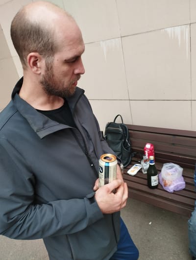 The image shows a man holding a can of beer. He is wearing a grey jacket and blue pants. In the background, there is a wooden bench with several items on it, including a black backpack, a carton of juice, a bottle of alcohol, and a plastic bag with food. The scene appears to be outdoors, possibly in a park or public space, with a tiled wall in the background. The man is looking to the right, and his expression is neutral. The overall impression is of a casual gathering or break. The city of Korolyov, Russia, is not explicitly visible in the image, but could be the setting for this scene.