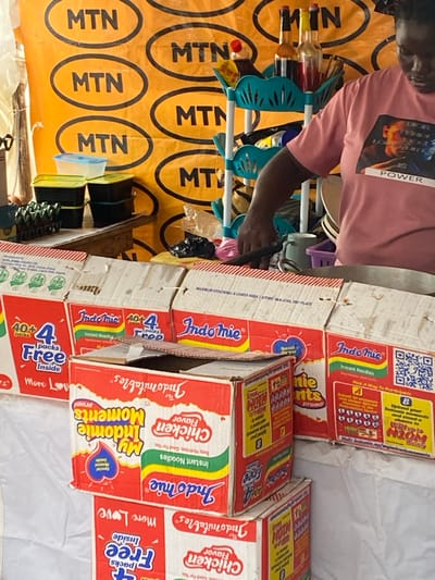 The image shows a market stall in Alagba, Nigeria, with a person in the foreground wearing a pink t-shirt with a graphic and the word "POWER" on it. They are standing behind a display of various products, primarily boxes of Indomie instant noodles. The background features a yellow banner with multiple black "MTN" logos.
The products are arranged in stacks and some are open, revealing packets of noodles. Some boxes highlight promotions such as "40+4 packs Free Inside" and "My Indomie Moments". The lighting suggests it could be daytime, with no specific weather conditions apparent. The overall scene depicts a commercial transaction in a local market setting. Visible text includes brand names like "MTN" and "Indomie," as well as promotional slogans and nutritional information on the product packaging. There are also some bottles of liquid and other smaller items on shelves to the left of the main display.