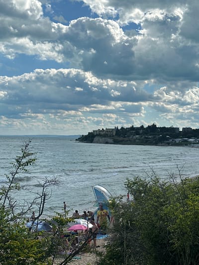 The image depicts a sunlit beach scene at Topola, Bulgaria. Several people are visible on the sandy shore, some relaxing under umbrellas and others wading or swimming in the gently rippling water. One person is wearing a yellow t-shirt. In the background, lush green trees line a cliff overlooking the sea. Above, dramatic clouds fill the sky, suggesting a partly cloudy day. The overall mood is relaxed and leisurely, capturing a typical summer day at the beach. The photo appears to have been taken during daylight hours. A blue and white umbrella with visible text is present on the beach.