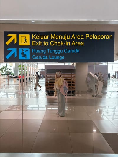 A person stands in front of a glass door at what appears to be an airport terminal. A sign above the door indicates directions in both Indonesian and English. The top line reads "Keluar Menuju Area Pelaporan" and "Exit to Check-in Area," with a yellow arrow pointing right and a yellow figure exiting a door. The second line reads "Ruang Tunggu Garuda" and "Garuda Lounge," with a blue arrow pointing right and a blue icon of a person seated next to an airplane. The person in the foreground is wearing a yellow top and light blue jeans and carrying a pink backpack. They are holding a phone and appear to be taking a selfie. Behind the glass, the terminal interior is visible, with seating areas, plants, and other travelers. The floor is tiled with a reflective surface, creating mirrored images.