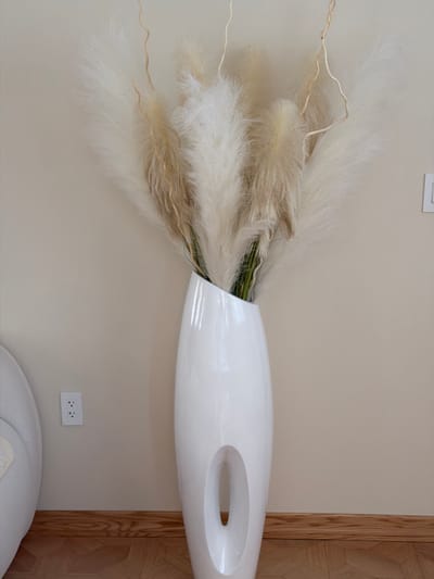 A tall, white, modern vase with an oval cutout sits on a hardwood floor, filled with a large arrangement of dried pampas grass and thin, wispy branches. The vase is positioned against a plain, beige wall. To the left of the vase, a portion of a white armchair is visible. On the wall to the left of the vase, an electrical outlet with two sockets is present. The scene is well-lit, suggesting an indoor environment. There are no people or animals visible in the image. The location is identified as Marlboro Township, United States of America, based on the provided context.