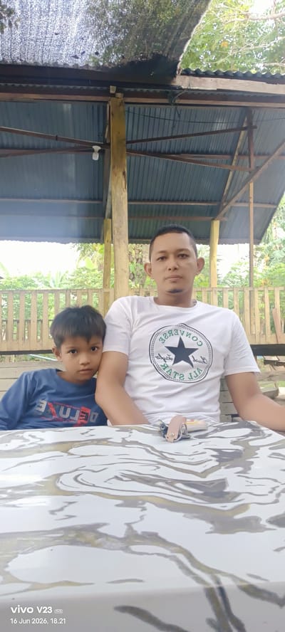In an outdoor, covered seating area with a corrugated metal roof supported by wooden beams and posts, a man and a young boy are seated at a table. The man, on the right, wears a white t-shirt with a black Converse logo. The boy, on the left, is dressed in a blue long-sleeved shirt with red lettering. They are positioned in front of a light-colored table with a marbled pattern. The background consists of greenery and a wooden fence. The scene is located in Glokaron, Indonesia.