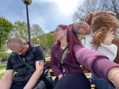 Three individuals are sitting on a park bench. A man with a shaved head and a black vest is sitting on the left, looking down. A woman with red hair and glasses is seated in the middle, holding what appears to be a pen or cigarette. To the right, a woman is holding a brown poodle with her eyes closed. The background shows greenery with trees, suggesting a park location. A lamppost is visible behind the man, and the scene appears to be taken in daylight under a sunny sky.
The photograph was taken in the city of Nikopol, Bulgaria.