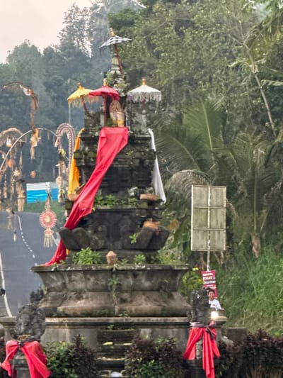 The image depicts an outdoor scene in Payangan, Indonesia. In the foreground, a stone structure, possibly a shrine or monument, is adorned with red fabric. To the left, a dark stone statue is also wrapped in red cloth. The middle ground features a larger, multi-tiered stone structure with several decorative umbrellas on top. A long red fabric drapes down the side of this structure. In the background, lush green vegetation and trees are visible. A road with white markings is on the left side of the image. There is a white rectangular object mounted on a pole, and a red banner with Indonesian text is visible. A person riding a white motorcycle is partially visible in the right background, looking towards the camera. The sky is overcast.