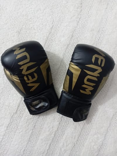 Two black and gold Venum boxing gloves are displayed on a textured white surface. The gloves are positioned diagonally, with the left glove slightly above and to the left of the right glove. Both gloves feature the "VENUM" logo printed in gold lettering. The left glove shows the "VENUM" logo vertically from top to bottom, while the right glove displays it in reverse, from bottom to top. The white surface appears to be a textile, possibly a rug or blanket, with a ridged texture. There are no people present in the image, and no discernible location in Douala, Cameroon is indicated.