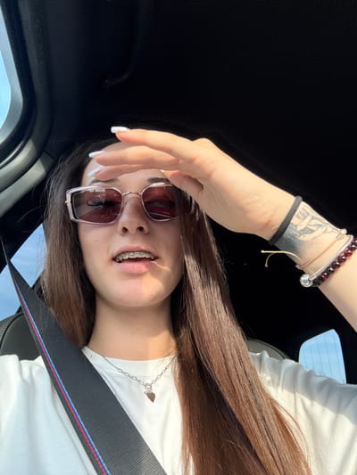 A young individual is seated in the passenger compartment of a vehicle, in the city of Ignatievo, Bulgaria. The subject, appearing female, has long brown hair and is wearing rectangular-framed sunglasses with pink/purple tinted lenses. Braces are visible on her lower teeth, and she has light freckles across her nose. Her fingernails are manicured with white tips. She is wearing a white short-sleeved t-shirt and a silver-colored chain necklace with a small heart-shaped pendant. Her left hand (viewer's left) is raised to her forehead, fingers slightly splayed, possibly shielding her eyes from direct sunlight. On her right wrist (viewer's right), a black hair tie is present, along with a black ink tattoo depicting an intricate, possibly knot-like or serpentine pattern. Two bracelets are also worn on this wrist: one thin string-like, and another beaded with dark red stones and a silver spherical charm. The interior of the vehicle is dark, with a black headliner. A black seatbelt, featuring prominent blue and red stripes, crosses the subject's torso. Outside the vehicle, a clear light blue sky is visible through the windows. The scene is brightly lit by natural daylight.