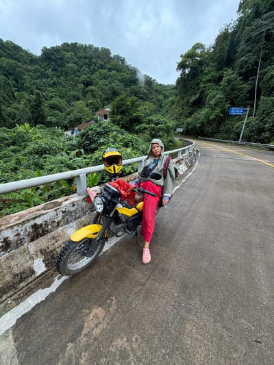 A person is sitting on a yellow motorcycle on the side of a road in a rural, mountainous area of Thượng Trạch Commune, Vietnam. The motorcycle is parked on a concrete bridge, with a metal railing to the left. The person is wearing a light-colored hooded jacket, red pants, and pink Crocs. A yellow helmet rests on the handlebars of the motorcycle, and a red bag is visible on the seat. The surrounding environment is lush and green, with dense vegetation and trees covering the hillsides. A road with yellow lines curves into the distance, and a blue sign with Vietnamese text is visible on the right side of the road. The sky is overcast, suggesting a cloudy or potentially rainy day.