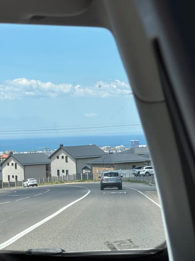 The image shows a view from inside a vehicle, looking out towards a coastal town and the sea. The perspective is slightly tilted, suggesting the photo was taken from a car window.
The foreground is dominated by the interior of the car, with the dark gray frame of the window and a lighter gray interior panel visible on the left and top. Beyond the window, a bright blue sky with scattered white clouds stretches across the scene.
In the middle ground, a road curves to the right. A silver car is visible on the road, driving away from the viewer. Further down the road, a white vehicle, possibly a van, is parked. Along the side of the road, a row of modern houses with gray roofs and light-colored walls are situated. Some of these houses have multiple windows visible.
In the background, the vast expanse of the blue sea meets the horizon. Beyond the sea, a cityscape with several tall buildings is visible in the distance. The overall impression is that of a sunny day in a coastal residential area. The lighting suggests it is daytime, likely mid-morning or afternoon. There is no visible text in the image.