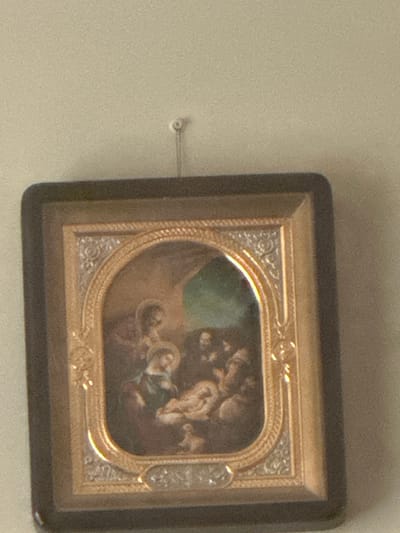 A framed painting of the Nativity scene is hanging on a textured, light-colored wall, possibly a wall in a home. The painting is oval-shaped with a dark, rectangular outer frame and a decorative gold inner frame. The artwork depicts a central figure of the Virgin Mary holding the infant Jesus in a manger. Joseph is seated beside them, and other figures appear to be present. The background suggests an indoor setting with an opening to a dimly lit green area. A small, white hook is visible on the wall above the painting. The image is blurry and taken at an angle, making it difficult to discern finer details of the painting and its immediate surroundings. The location in Yavoriv, Ukraine, is not visually evident from the provided image.