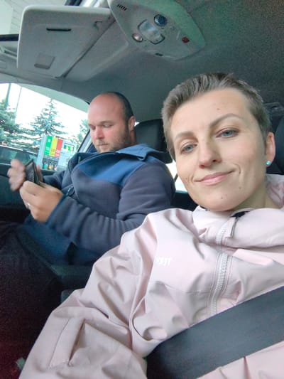 Two individuals, a man and a woman, are seated inside a car. The man, with a bald head and beard, is in the driver's seat, looking down at a wallet in his hands. The woman, with short hair, is in the passenger seat, smiling and looking towards the camera. They appear to be at a gas station, with gas pumps visible through the car window. The lighting suggests it is daytime. The car interior includes a sun visor with a light fixture. The woman is wearing a light pink jacket. The man is wearing a dark blue and grey jacket.