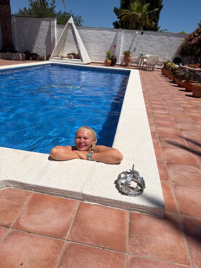 In Mont-roig del Camp, Spain, a woman with blonde hair pulled into braids is partially submerged in a bright blue swimming pool, leaning on the pool's edge. She is wearing a watch on her left wrist and a turquoise pendant on a chain. Next to her, on the pool's edge, is a small, ornate silver crown with leaf motifs. The pool is surrounded by a red tiled patio. In the background, a white stucco wall encloses the property, with a triangular white cabana containing cushions and a rolled-up mat. Various potted plants, including cacti, and patio furniture are visible along the wall. The sky is clear and blue, indicating a sunny day.