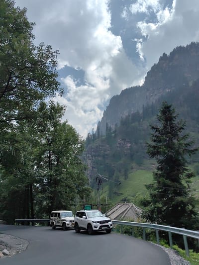 In Kothi, India, two white SUVs are on a paved road that curves to the right, bordered by a metal guardrail. The SUV in the foreground is white and has a license plate that reads "HP 33G 5808". Behind it is another white SUV, a Mahindra Bolero. Tall green trees flank the left side of the road, and a large, rocky mountain covered in trees dominates the background. The sky is filled with dramatic, white clouds.