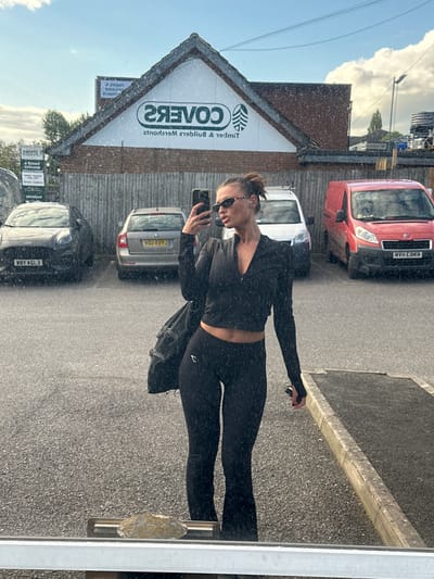 This image, rotated 90 degrees clockwise, presents a mirror reflection of a person taking a selfie in an outdoor commercial setting during bright daylight. The main subject is a person, reflected upright, holding a black smartphone to take the photo. They are dressed in black long-sleeved activewear, including a crop top that reveals their midriff and matching leggings with a small white logo on the upper left thigh. Dark sunglasses obscure their eyes, and their hair is tied back.
The background, also reflected, shows a paved area, likely a parking lot or street, consistent with the Tonbridge and Malling, United Kingdom context provided. Several vehicles are visible: a red van with the reversed license plate "KHE3 HAM", a white van, a silver hatchback displaying the reversed plate "TAJA CDA", and a black car with the reversed plate "WBY ASL3". A prominent building with a pointed roof is reflected in the upper left, featuring large, reversed text on its white facade that reads "COVERS" and "Timber & builders Merchants". The sky above is bright blue with visible white clouds, indicating sunny weather. The mirror surface has numerous small speckles, possibly dust or water.