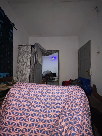 The image depicts the interior of a dimly lit room in Ilishan, Nigeria, viewed from a low perspective, likely from a bed. The foreground is dominated by a bed covered with a light pink/orange sheet featuring a repetitive blue geometric pattern of intersecting lines and stylized leaf motifs.
To the left, a dark blue curtain with light "CHANEL" logos hangs partially. Below it, several pairs of shoes, including light brown sneakers, rest on a surface. Adjacent to this, a tall, rectangular cabinet or wardrobe with a grey and white marble-like pattern stands. A light grey fabric and other clothes, including a striped item, are draped over the cabinet's top edge.
In the center, a doorway opens into an adjacent room. A black standing fan is positioned in this doorway, its circular cage visible. Inside the further room, a bluish light source hangs from the ceiling, and a dark rectangular object, possibly a television, is visible on a surface in the background. The floor in the second room appears to be tiled.
To the right of the main room, a closed grey door with a light-colored handle is present. Near the bottom right corner, a dark blue bag, possibly a duffel bag, is placed on the floor alongside some red fabric. A bare human foot, likely the right foot, is visible in the extreme lower right, resting on a dark surface next to the bed. The walls are light-colored, showing some discoloration and minor imperfections, particularly near the ceiling, which also exhibits some marks. No direct human interaction is depicted, but the presence of the foot suggests an individual is within the immediate vicinity.