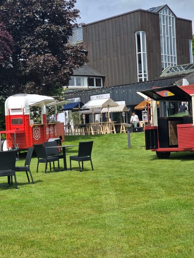 The scene depicts a food stand or bar setup on a well-maintained grassy lawn, likely in Lenningen, Luxembourg. A distinctive modern building with dark wooden cladding and geometric windows stands in the background. The setup includes a red and white mobile food stall, several black chairs, and tables. There are people visible within and near the stall. The setting suggests an outdoor event or market in a relatively green and open area. The scene is bathed in sunlight.