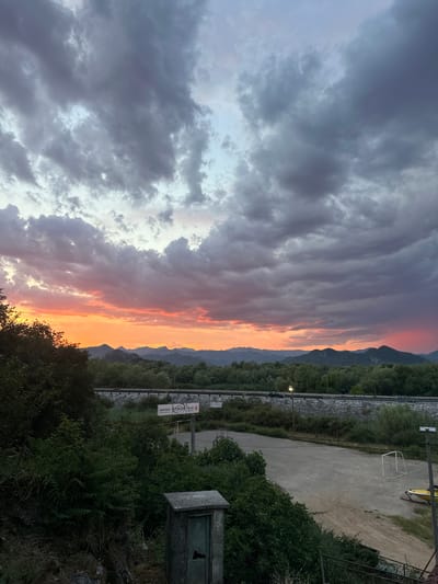 This is a vertical, wide-angle photograph of a sunset over a mountainous landscape. The sky is filled with dramatic clouds, transitioning from dark gray on the left to vibrant orange and pink hues on the right, illuminated by the setting sun. A mountain range is visible in the distance, silhouetted against the colorful sky.
In the foreground and midground, there is lush green vegetation, including trees and bushes. A paved area, possibly a sports court or parking lot, is visible with a small white structure, resembling a goalpost, and a yellow object that could be a vehicle or piece of equipment. A fence runs along the edge of the paved area, separating it from the surrounding vegetation. There are also a few signs with text, but they are too small and blurry to be read clearly. One sign appears to have red and white lettering. A small, weathered, green electrical or utility box is situated on the right side of the frame. The scene suggests an outdoor recreational area in a natural setting.
The time of day is clearly sunset, indicated by the colorful sky and the low angle of the light. The weather appears to be partly cloudy, contributing to the dramatic sky. The location is Vranjina, Montenegro, as indicated by the context. The overall mood is serene and awe-inspiring, with the natural beauty of the sunset being the dominant element.