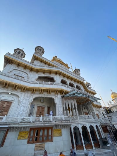 This image captures an exterior view of the Golden Temple in Amritsar, India, under a clear blue sky. The ornate white marble structure, adorned with gold accents, is the central subject. The building features multiple levels with balconies, arches, and domed turrets. A prominent clock is visible on the facade. Several individuals are present in the foreground, seated or standing, some wearing traditional attire. The overall environment suggests a public religious site, likely during daylight hours given the clear sky and shadows.